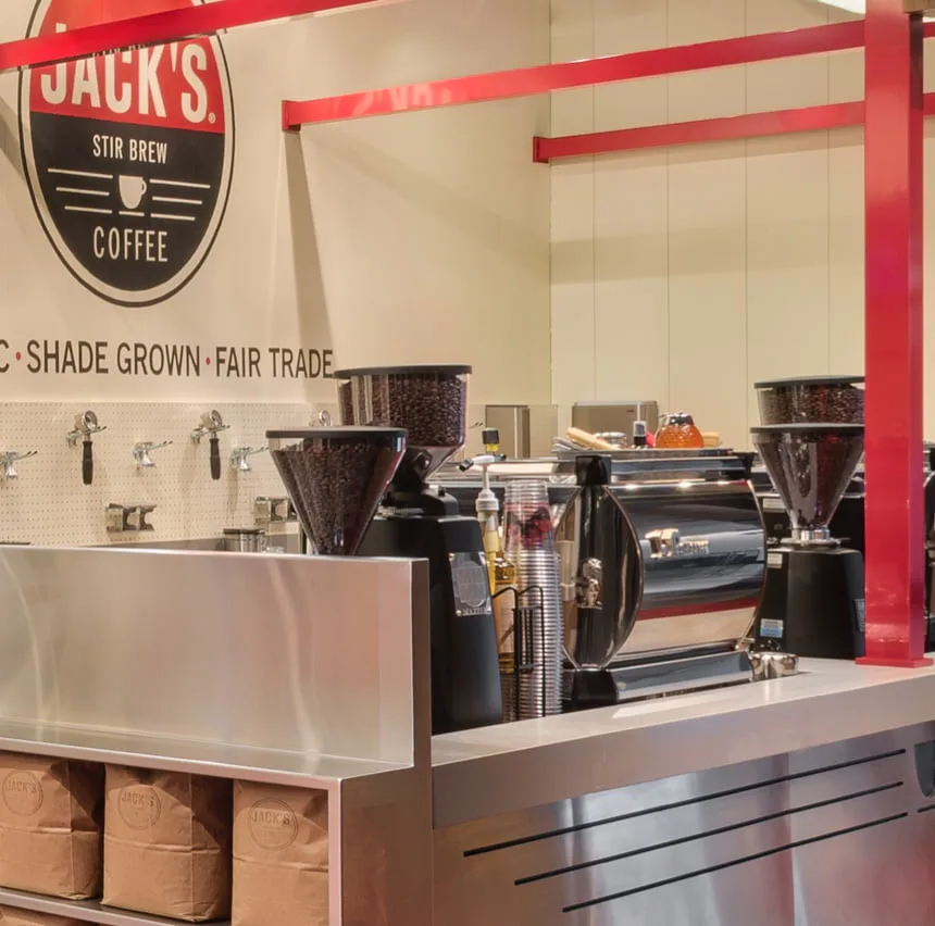 Modern coffee shop counter with black espresso machines, white counter front, and red architectural beams, designed by EunHea Kim.