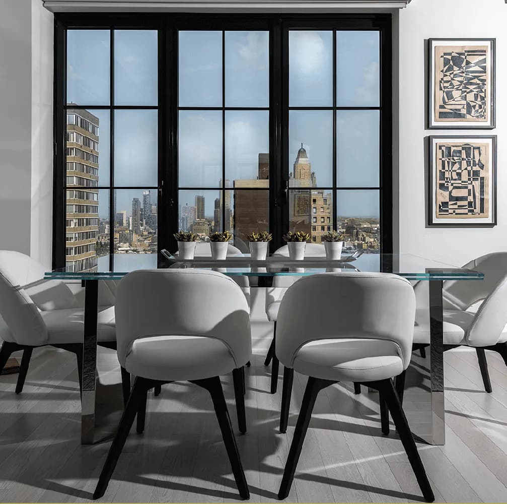 Modern dining space with gray upholstered chairs, glass table, and expansive black-framed windows overlooking a city skyline, designed by Kim Radovich.