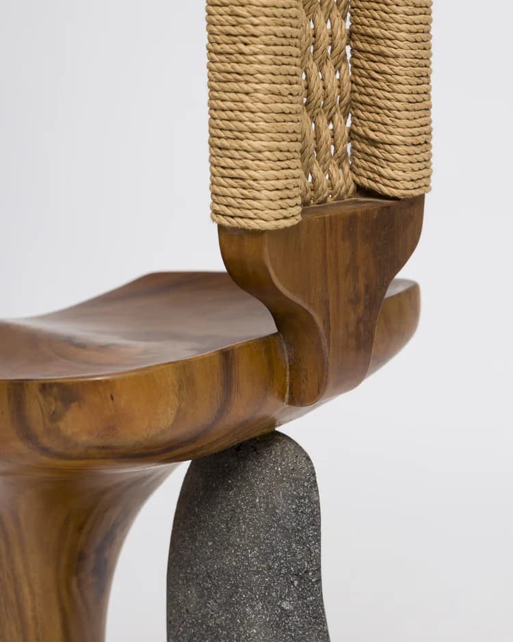 Close-up detail of a wooden sculptural chair or stool featuring curved organic forms, natural wood finish, rope-wrapped elements, and a dark stone base, by Kim Mupangilai.