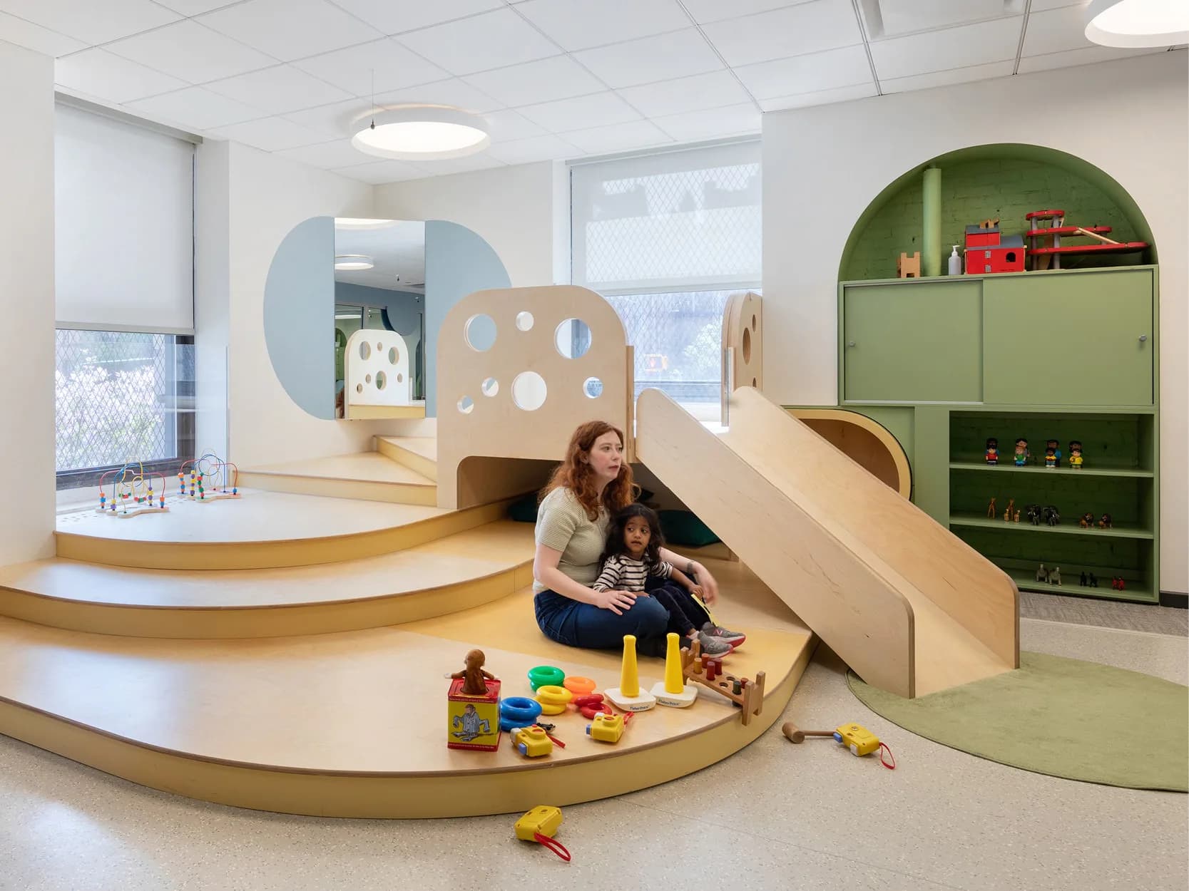 Curved cream and wood play area with integrated slide and storage, green cabinetry, and natural light in a modern childcare interior by Kim Yao.