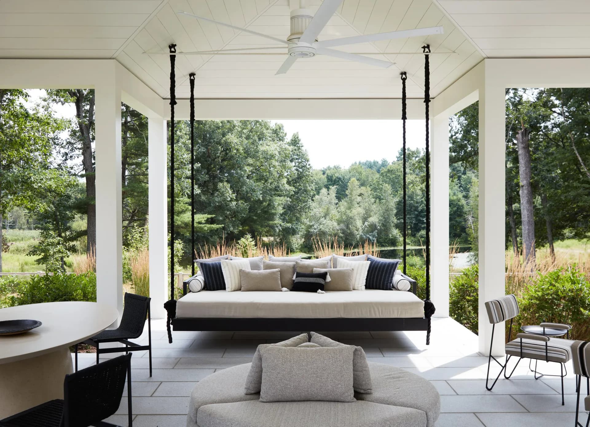 Modern bedroom pavilion with black four-poster bed, neutral linens, and expansive covered porch overlooking manicured grounds by Kirsten Flanigan.