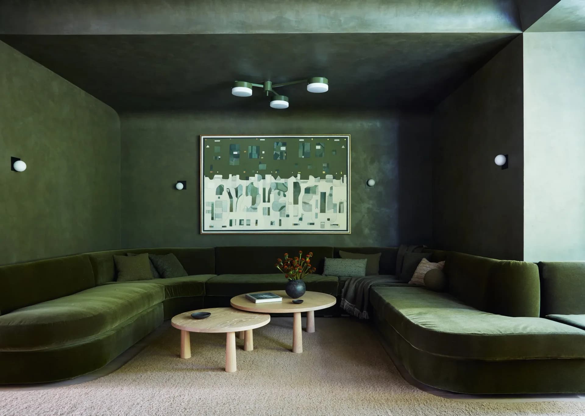 A sunken lounge with curved olive-green seating, natural wood tables, and abstract artwork, designed by Kirsten Flanigan.