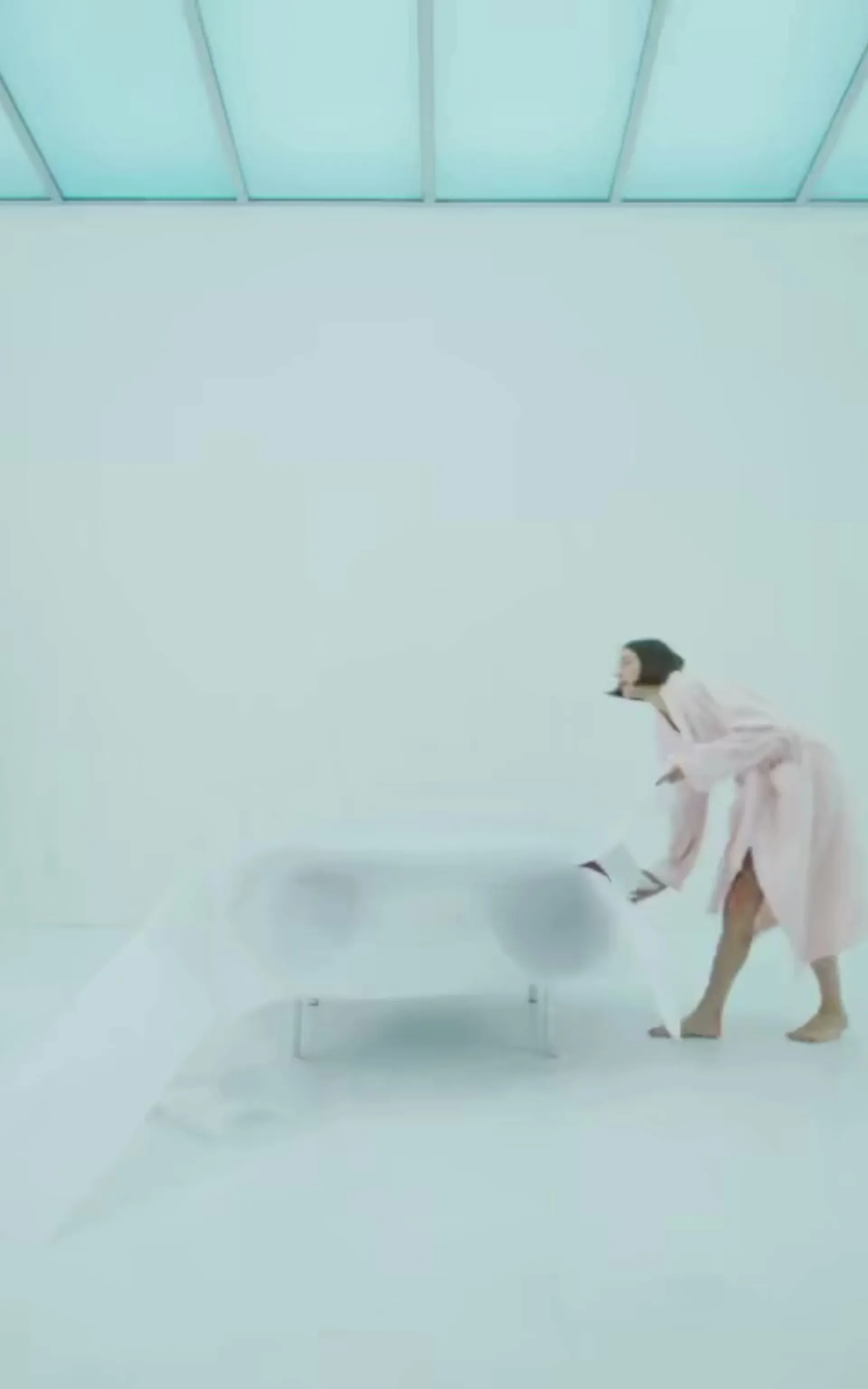 Minimalist white interior space with skylight ceiling and a person in pink coat beside a blurred sculptural form, by Kouros Maghsoudi.