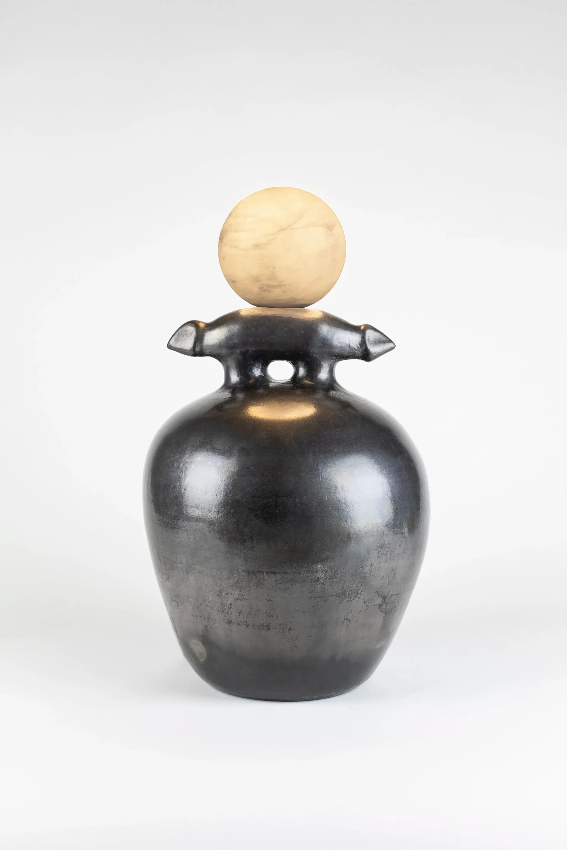 Sculptural vessel in dark polished metal with organic arm-like protrusions and a cream-colored spherical finial, creating a balanced composition by Kristin Victoria Barron.