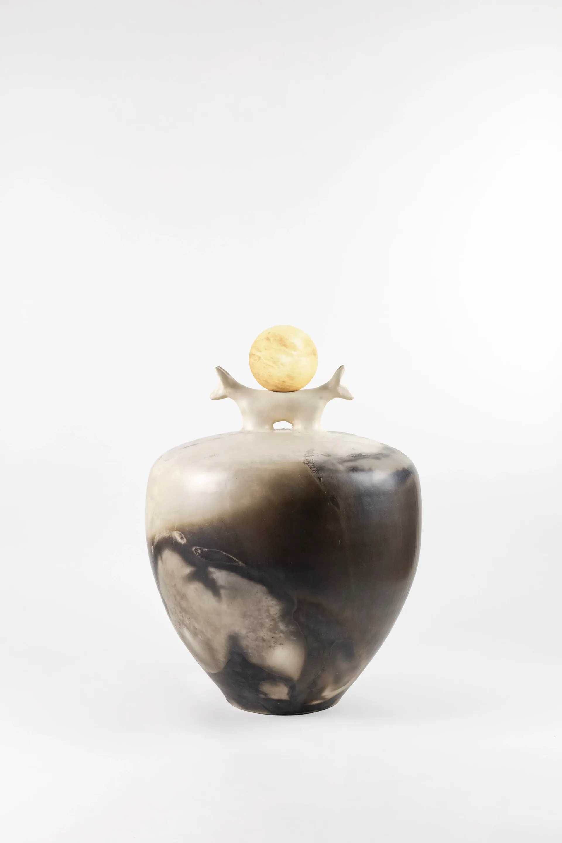 A sculptural ceramic vessel with dark marble-like glazing topped with a pale yellow sphere and abstract architectural finial, by Kristin Victoria Barron.