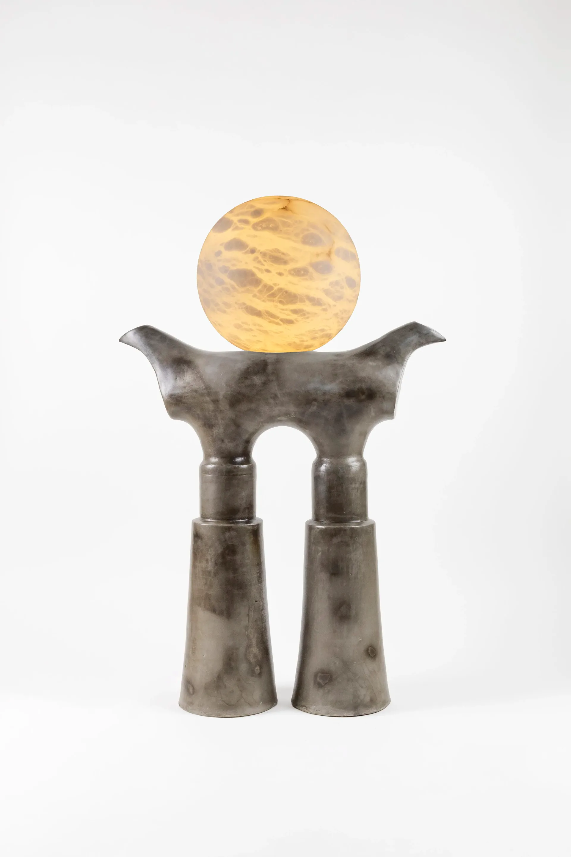 A sculptural installation featuring a dark metallic figure with upraised arms supporting a glowing yellow orb, set against a white background by Kristin Victoria Barron.