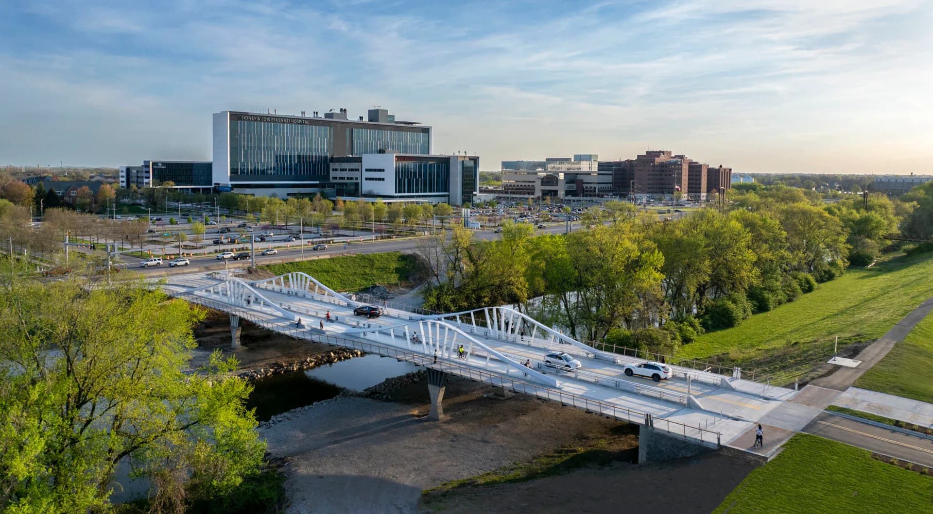 Modern white pedestrian bridge spanning a river with green landscaping, modern institutional buildings, and parking areas visible in the background by Kutay Biberoglu.