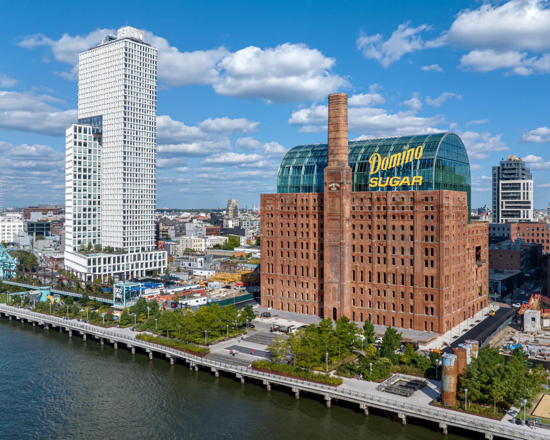 Waterfront view of a historic red brick building with green roofing and chimney, adjacent to modern white residential tower along the water in New York by Kutay Biberoglu.