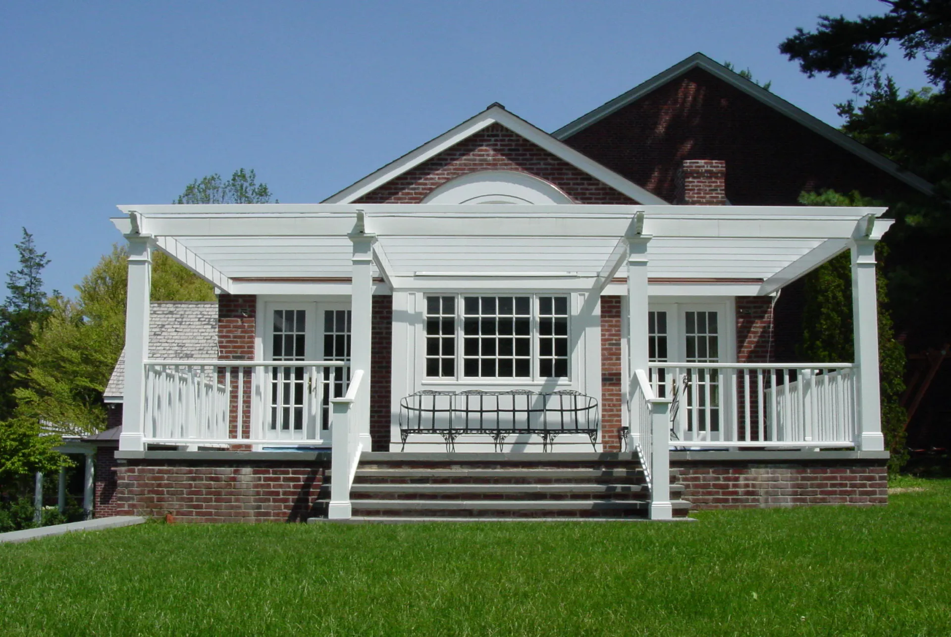 Modern white pergola-covered porch addition with brick foundation and large windows on residential home with manicured lawn, designed by Lainie Gahagan.