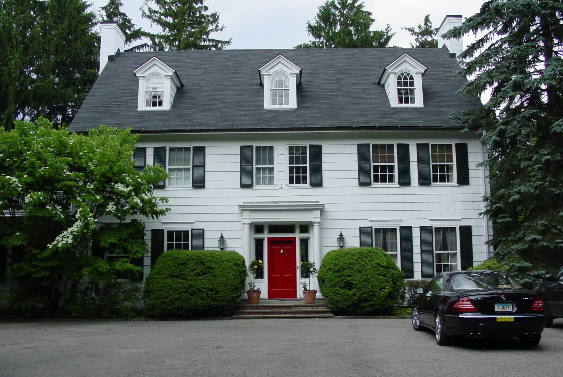 White colonial-style house with gray roof, red front door, black shutters, and manicured hedges, designed by Lainie Gahagan.