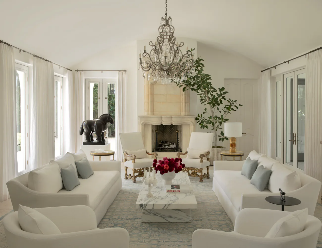Bright, symmetrical living room with cream curved sofas, fireplace, crystal chandelier, and expansive windows by Lauren Bensmiller.