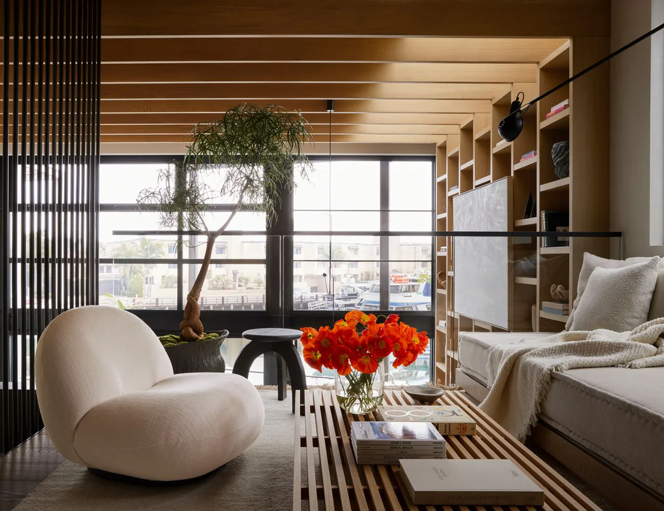 Modern residential interior featuring a sculptural cream armchair, wooden shelving, large windows overlooking a city, and vibrant orange floral arrangement by Lauren Bensmiller.