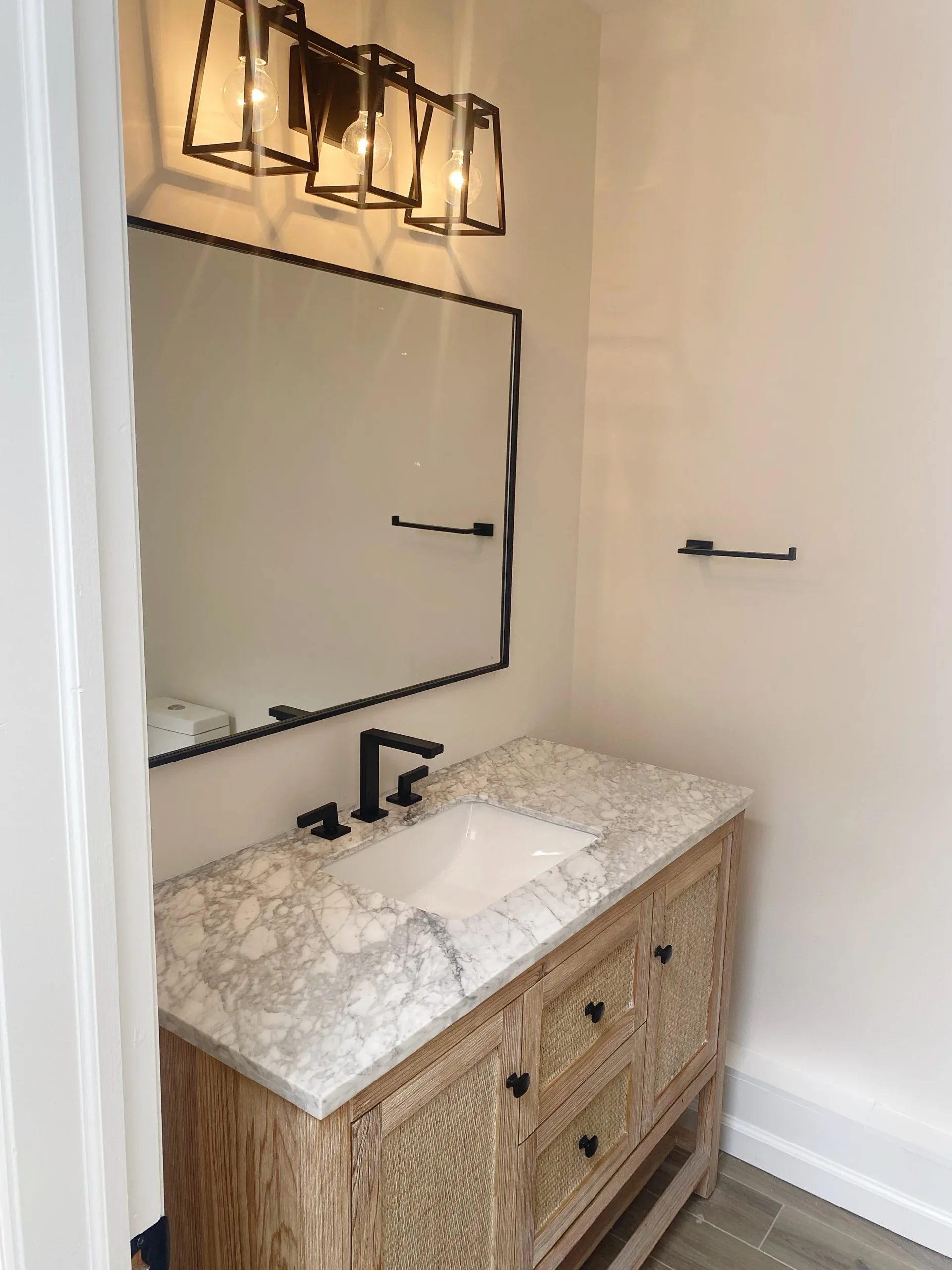 Modern bathroom vanity with marble countertop, wooden cabinetry, black fixtures, and framed mirror with sconces by Lauren Farhi.