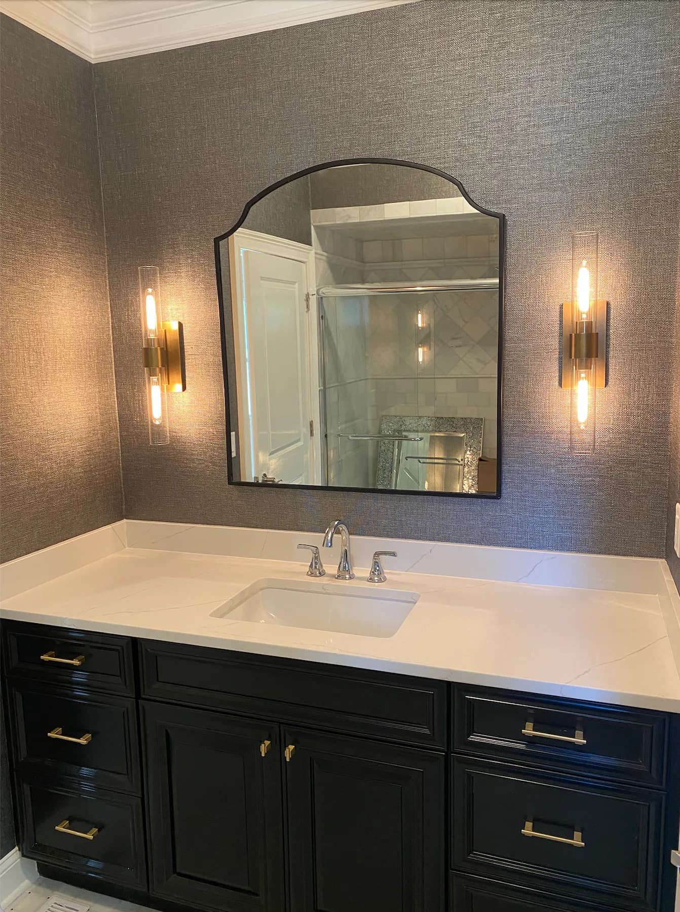 Modern bathroom vanity with black cabinetry, white countertop, arched mirror with black frame, and symmetrical brass sconces on textured gray walls by Lauren Farhi.