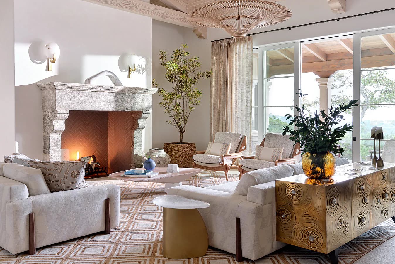 Elegant living room with cream upholstered seating, marble fireplace, brass accents, and large windows overlooking gardens by Leah Amelung.