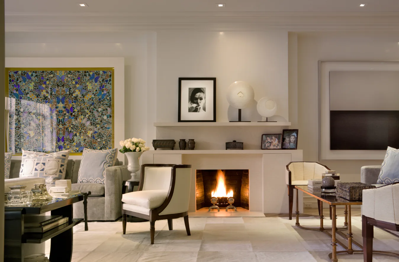 Luxurious living room with cream and beige palette, gas fireplace, built-in shelving, upholstered armchairs, and large window with blue patterned curtains by Leah Amelung.