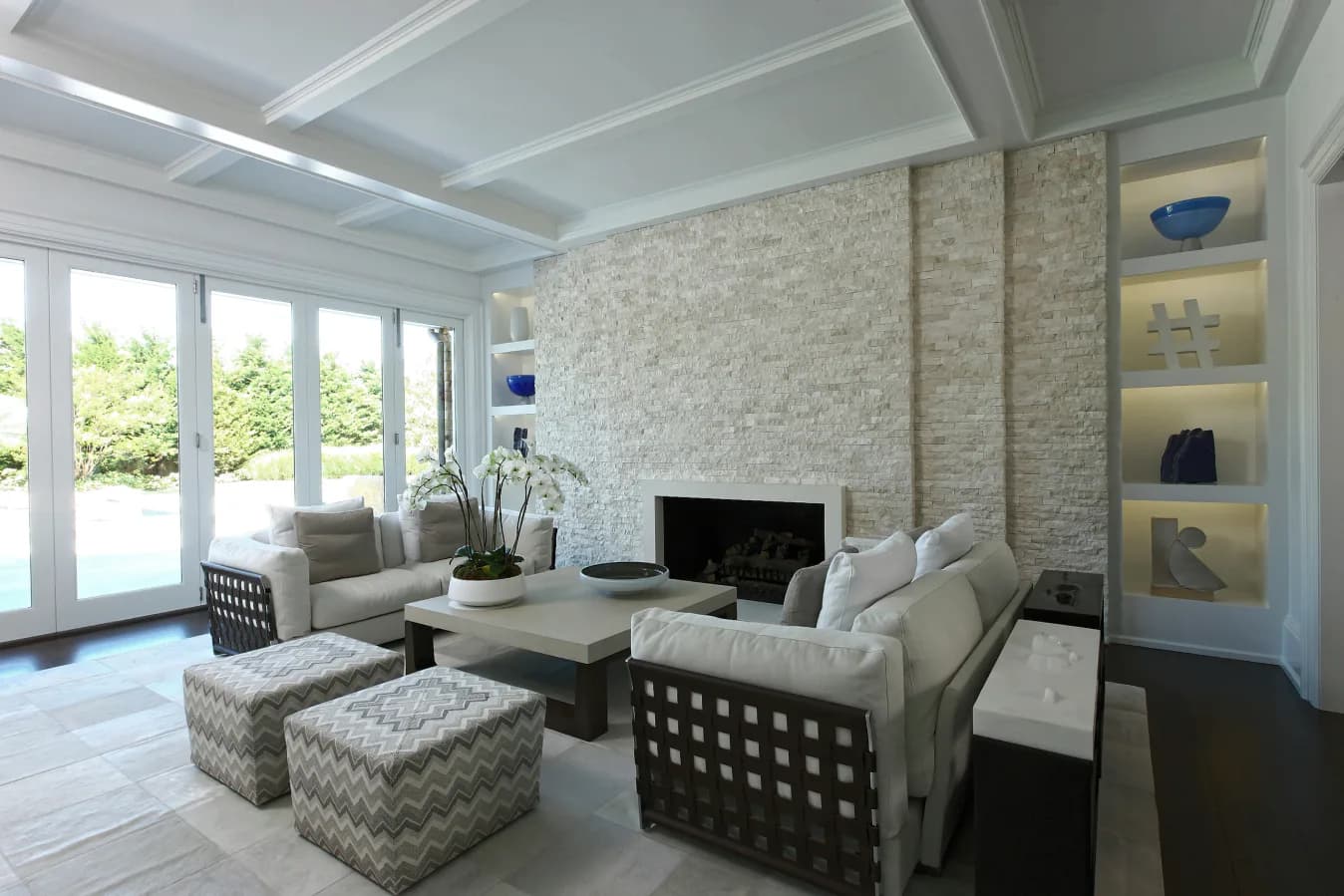 Modern living room with coffered ceiling, stone accent wall, cream seating, dark wood frame chair, and built-in shelving by Leah Amelung.