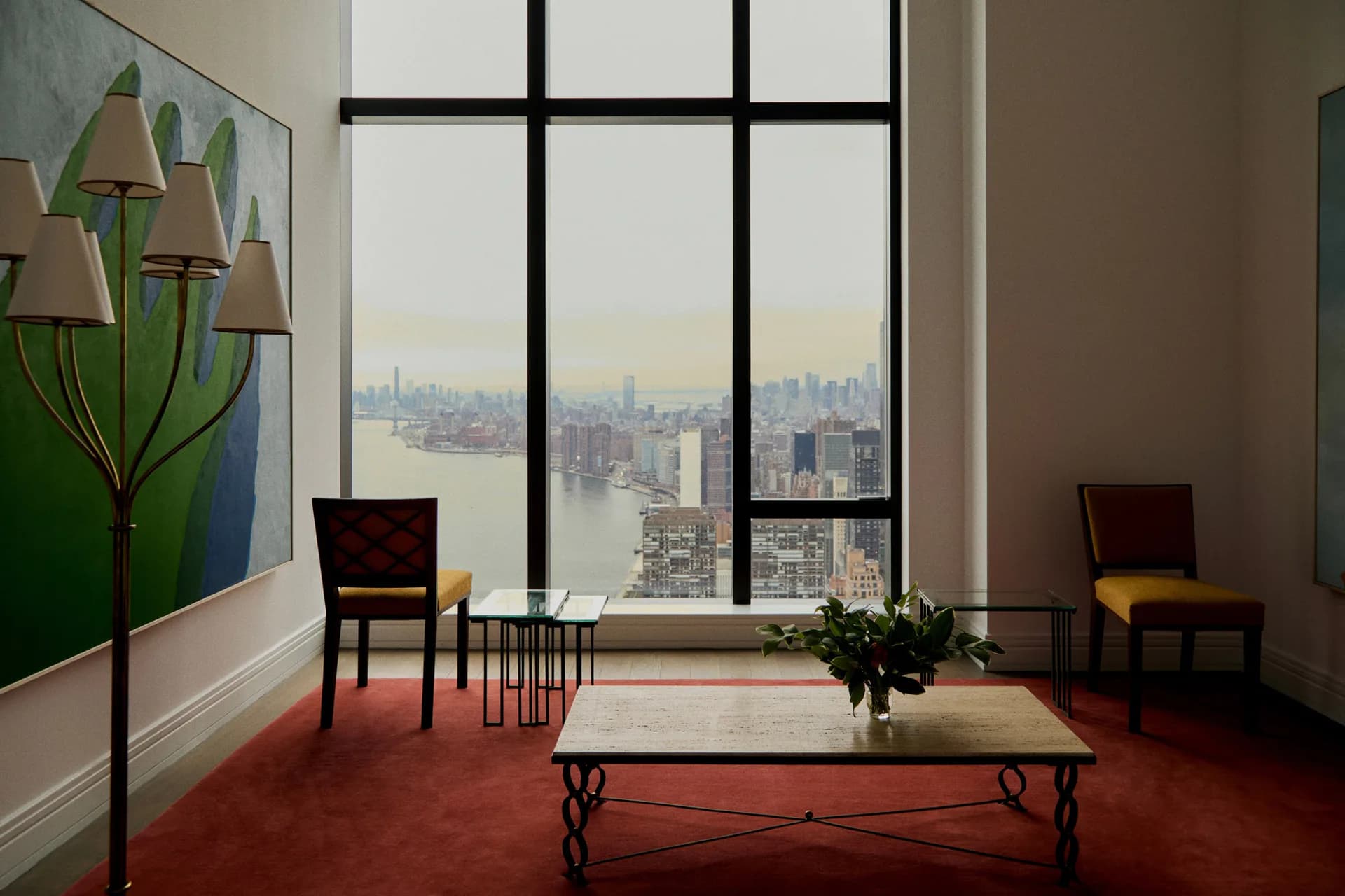High-rise interior with floor-to-ceiling windows overlooking Manhattan, featuring red carpet, modernist furniture, artwork, and potted plant by Lena Schipper.