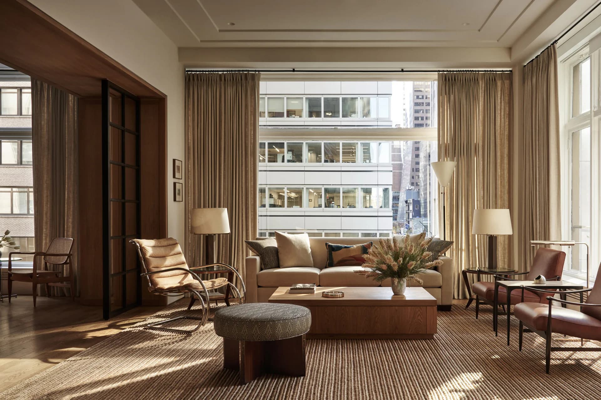 Luxurious Manhattan living room with floor-to-ceiling windows, neutral taupe furnishings, mid-century modern chairs, and natural light streaming across striped area rug by Lena Schipper.