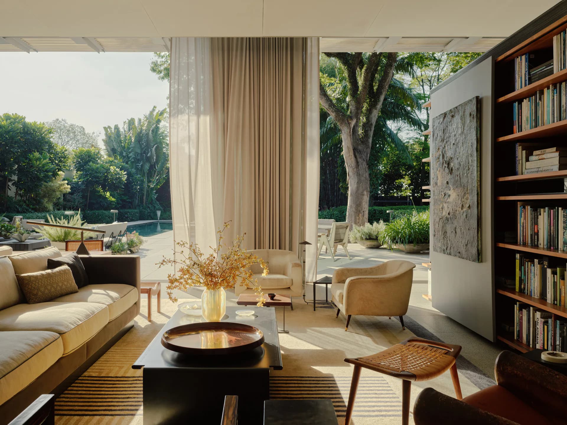 Sunlit modern living space with golden sofa, circular fireplace, bookshelf, and garden views framed by floor-to-ceiling curtains by Lena Schipper.