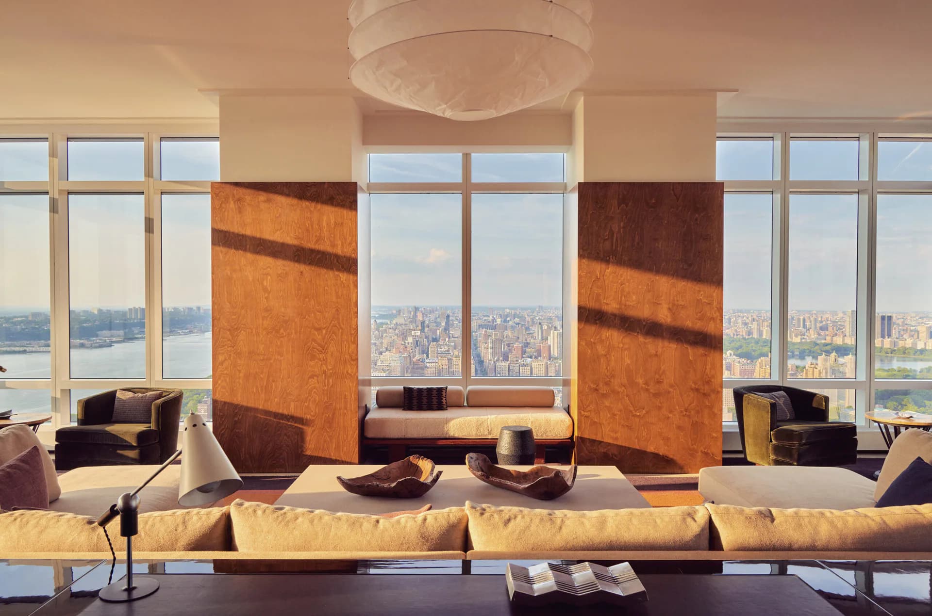 High-rise lounge interior with floor-to-ceiling windows overlooking a cityscape, featuring burnt-orange sculptural wall panels, modernist seating, and warm ambient lighting by Lena Schipper.