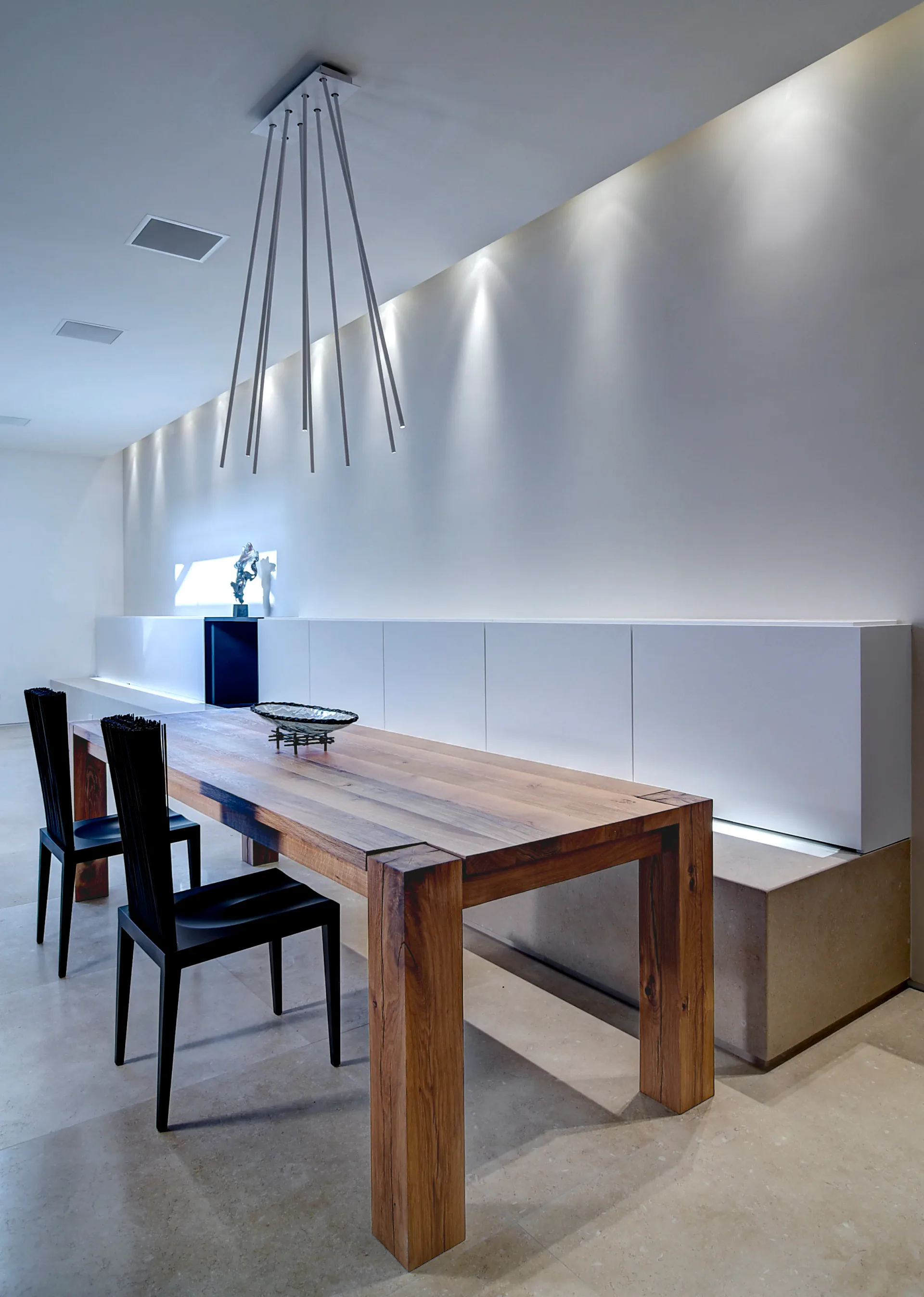 Minimalist dining space with natural wood table, black chairs, pendant lighting fixture, and white paneled wall with subtle accent lighting by Leopoldo Rosati.