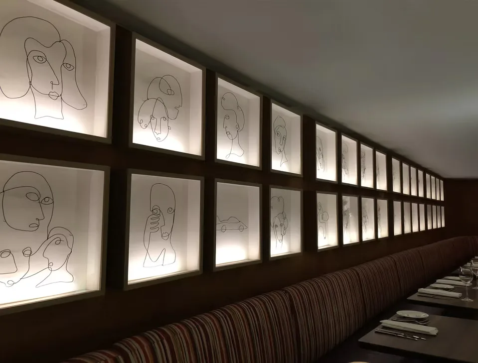 Hallway with illuminated wall panels displaying minimalist line-drawn portraits in a grid formation above upholstered seating by Leslie Ann Wigon.