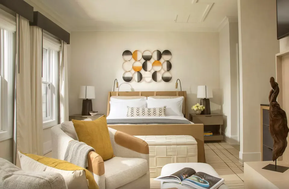 Serene bedroom with warm wood bed frame, cream and gold palette, geometric wall art, and natural light from windows by Leslie Ann Wigon.