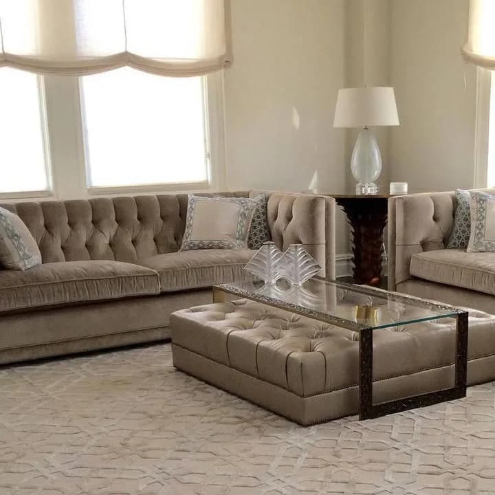 Luxe neutral living room featuring tufted sectional sofa, ottoman coffee table with glass top, decorative pillows, and classic table lamp by Leslie Weiss.
