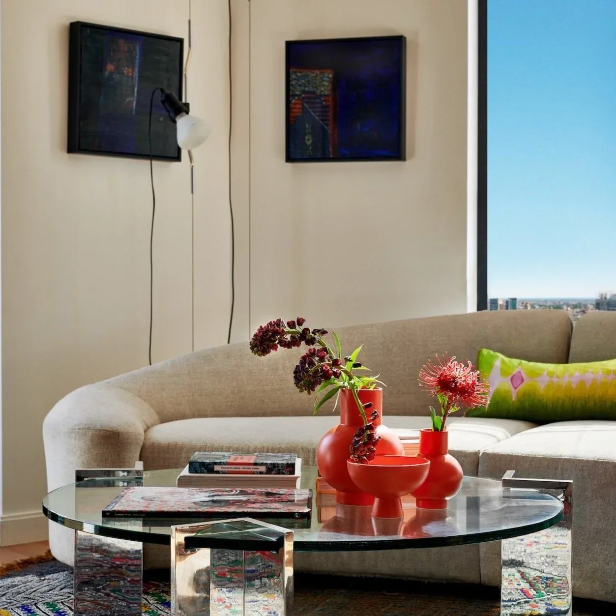 Modern living room with curved cream sofa, glass-top oval coffee table, red sculptural vase with flowers, and city views, designed by Leyden Lewis.