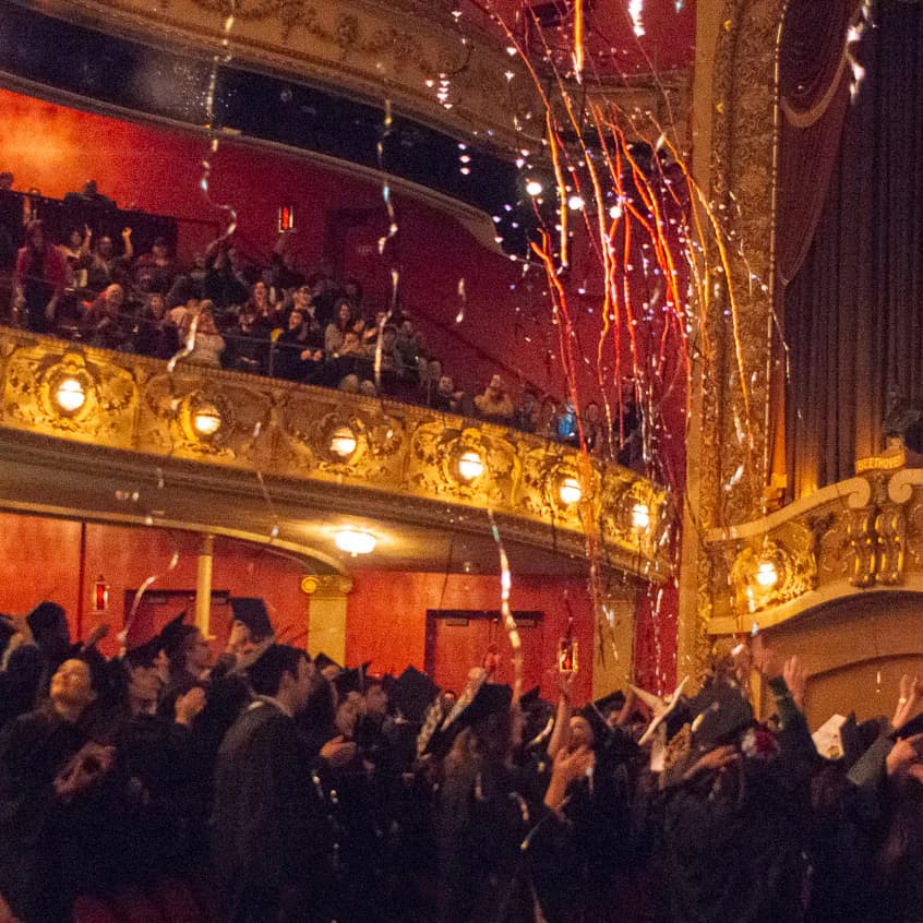 Ornate historic theater interior with gilt balcony railings, warm amber uplighting, and red velvet walls filled with evening guests by Lilly Roskopf.