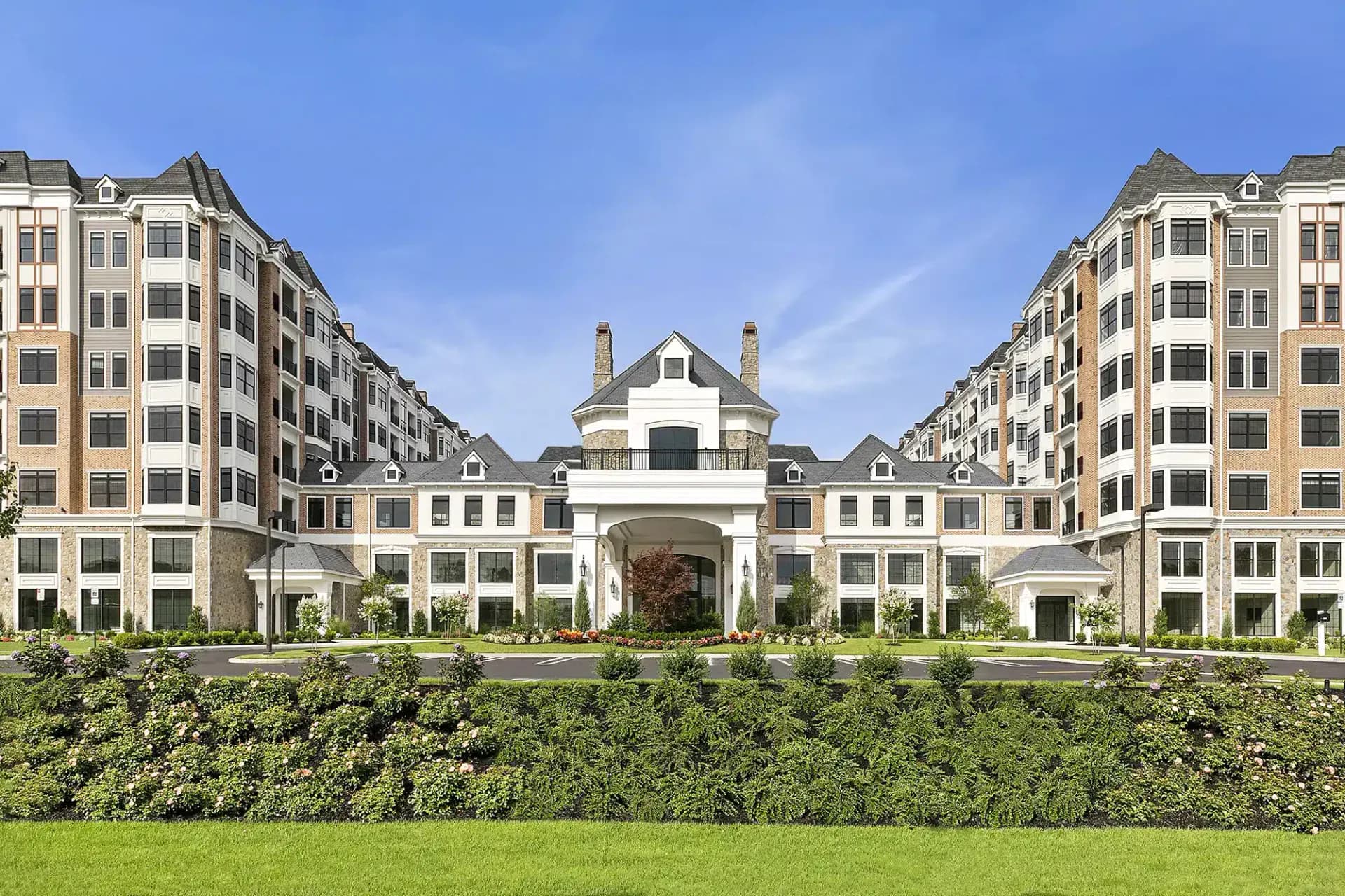 Architectural rendering of a symmetrical residential complex with cream and tan facade, central gatehouse with cupola, flanked by multi-story apartment buildings, manicured hedges and lawn by Lindsay Fauth.