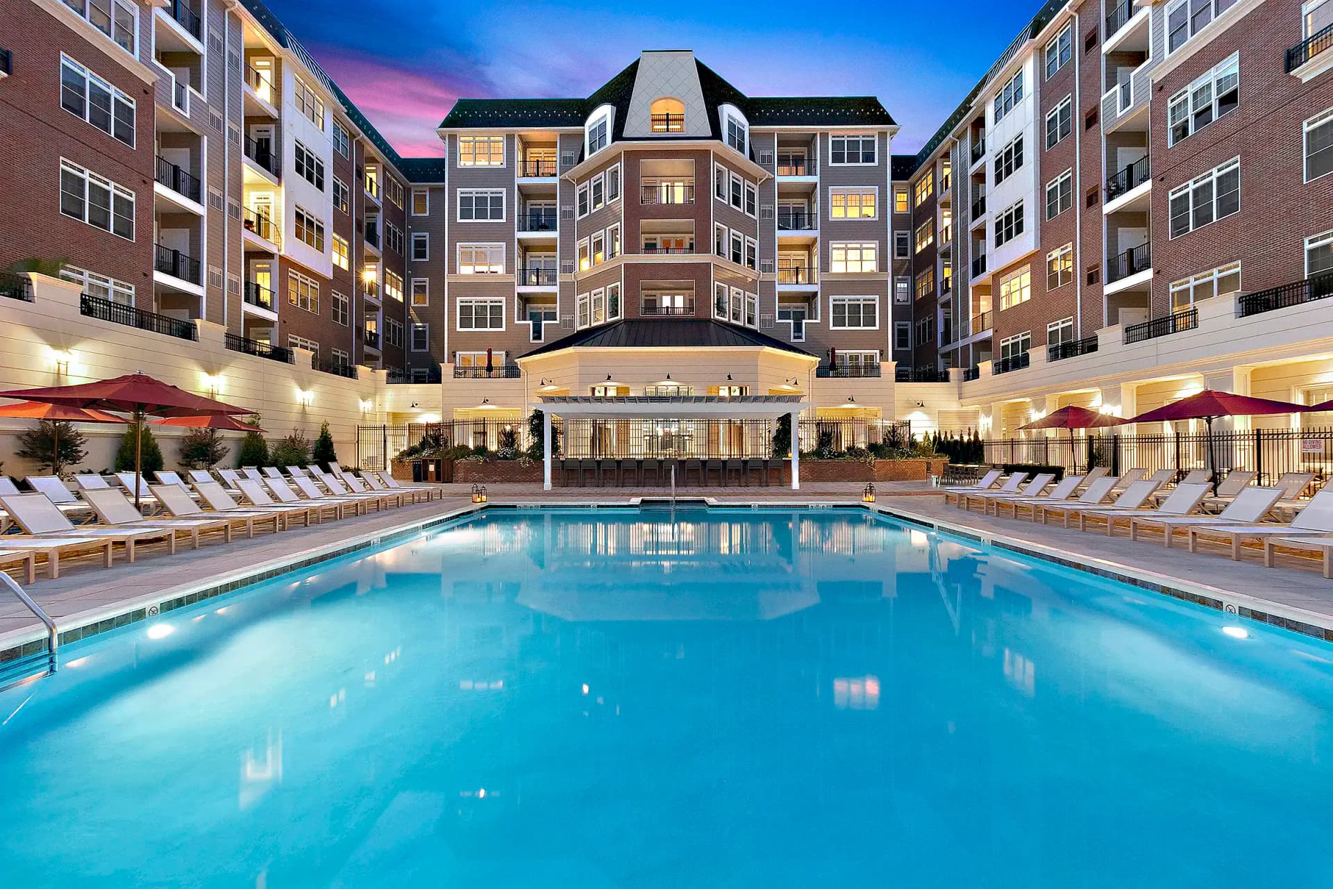 Twilight rendering of a luxury residential complex courtyard with illuminated pool, lounge seating, and multi-story brick buildings by Lindsay Fauth.
