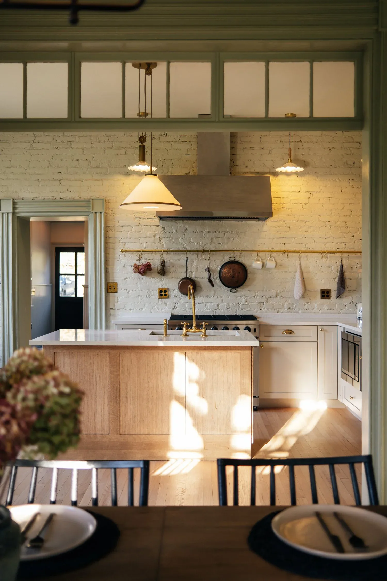 A bright, minimalist kitchen with white brick walls, natural wood cabinetry, pendant lighting, and open shelving displaying cookware, designed by Linnea Zeiloth.