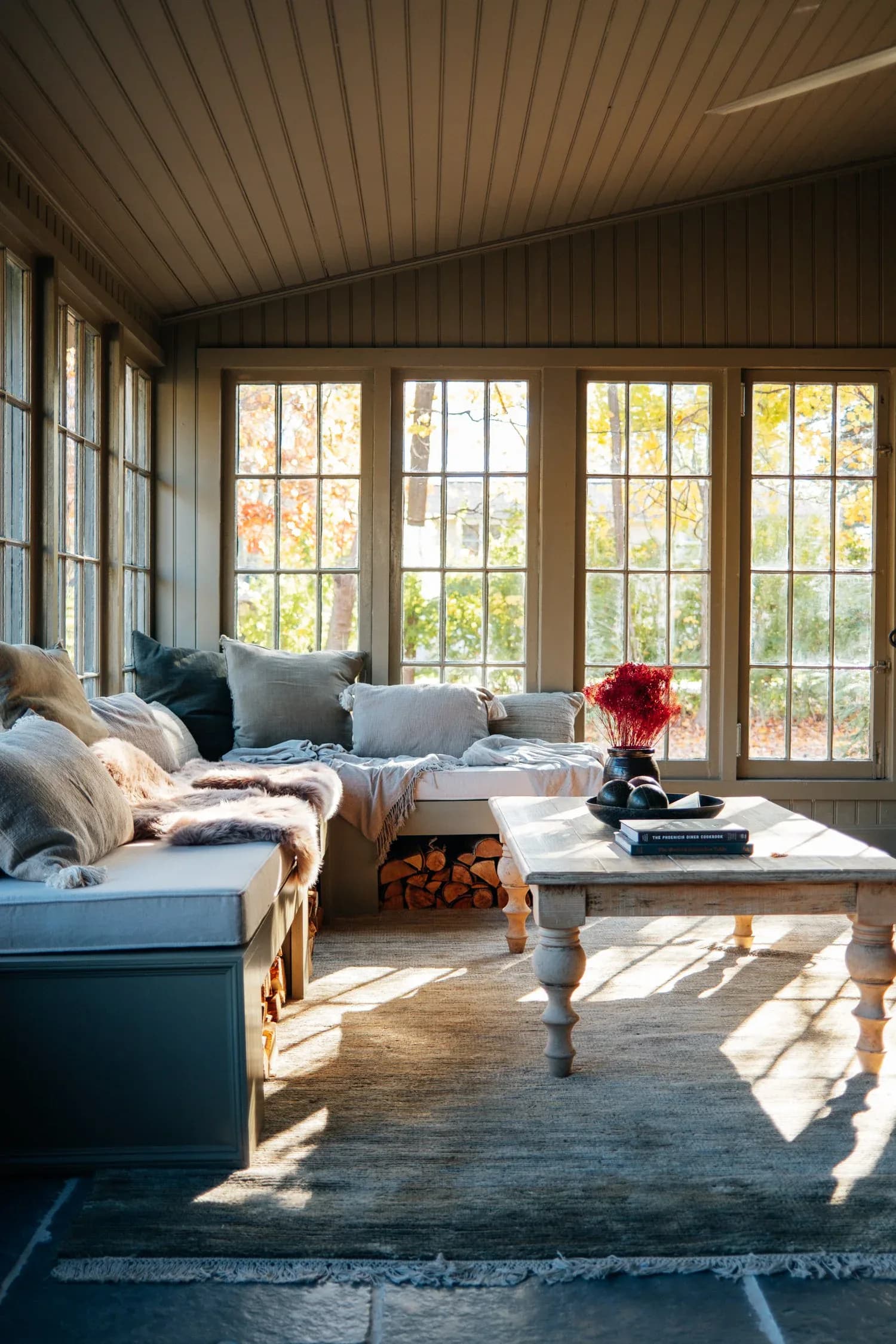 Bright sunroom with expansive gridded windows, blue upholstered seating, white coffee table, and natural wood ceiling by Linnea Zeiloth.