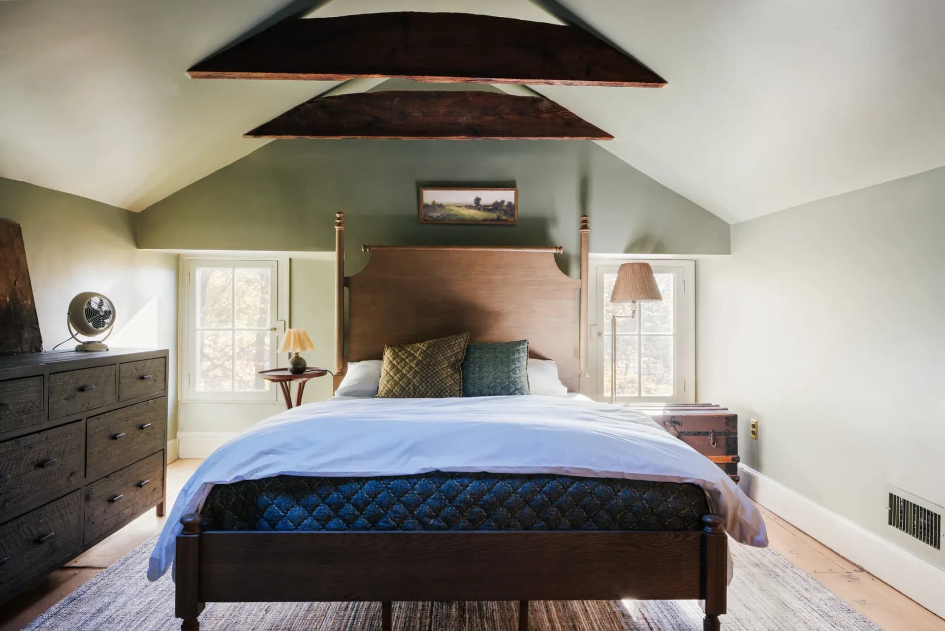 A bright bedroom with vaulted ceilings, sage green walls, dark wood ceiling beams, and a black metal bed frame, designed by Linnea Zeiloth.
