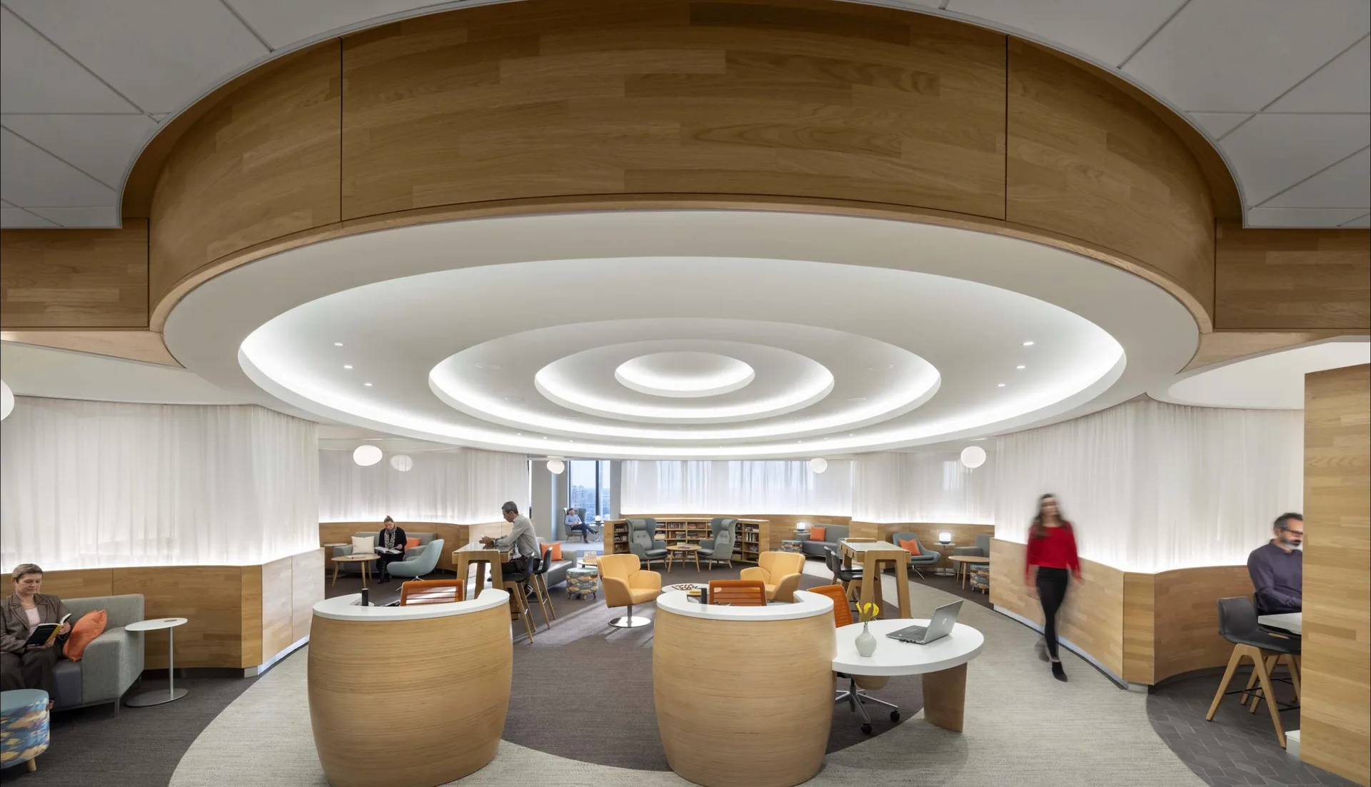 Modern open-plan office with curved wooden ceiling elements, concentric circular light rings, natural wood desks, and workspaces designed by Lionel Ohayon.