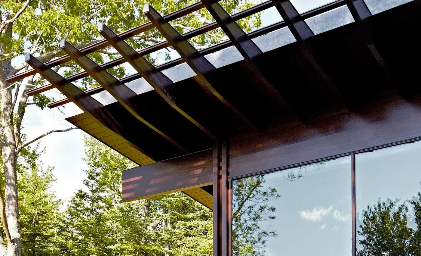 Modern architectural exterior featuring a dark steel pergola with lattice overhead structure, floor-to-ceiling reflective windows, and integrated natural greenery by Lisa Dang.
