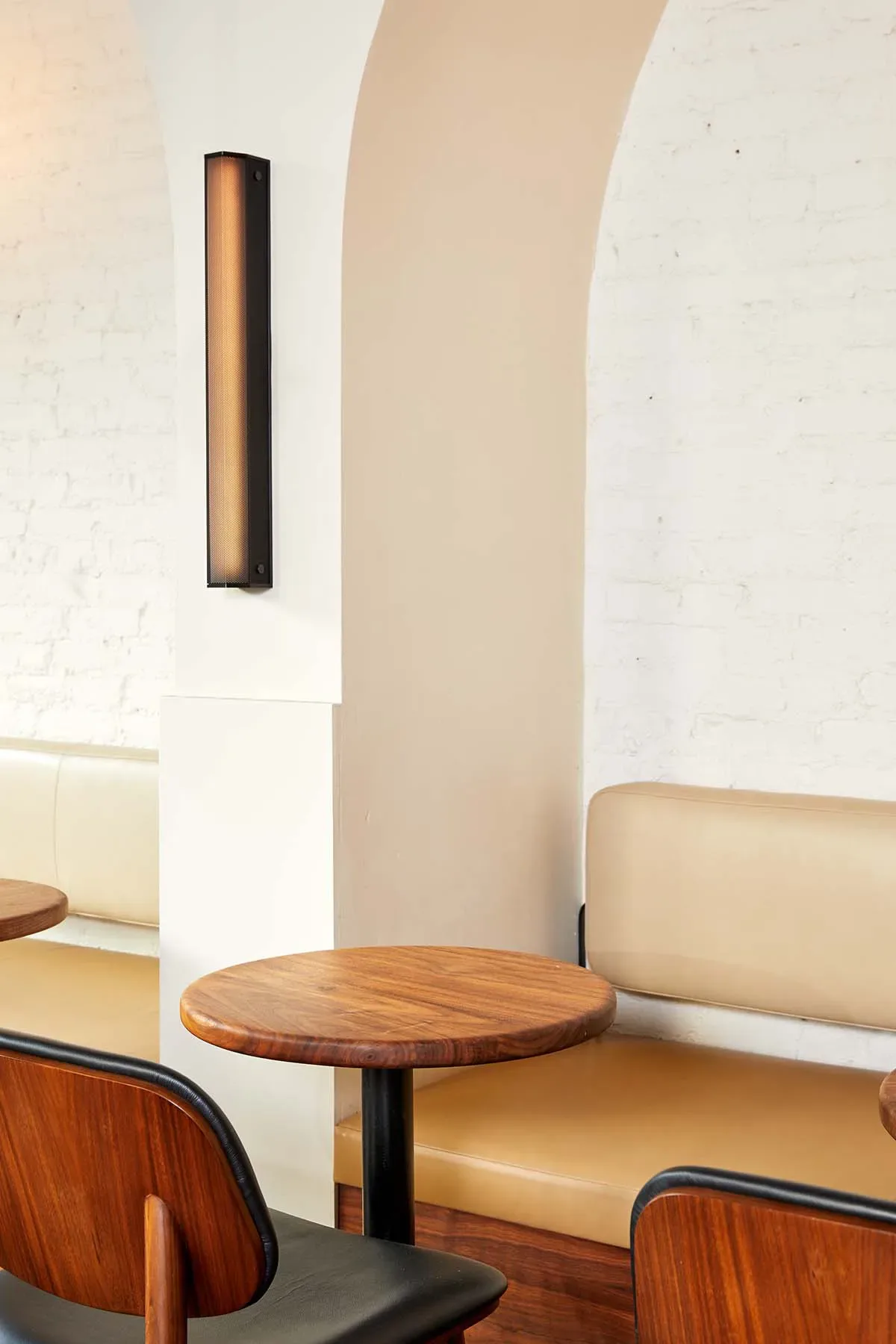 Modern lounge seating area with tan leather banquette, wooden round table, and minimalist linear wall sconce lighting by Lisa Galano.