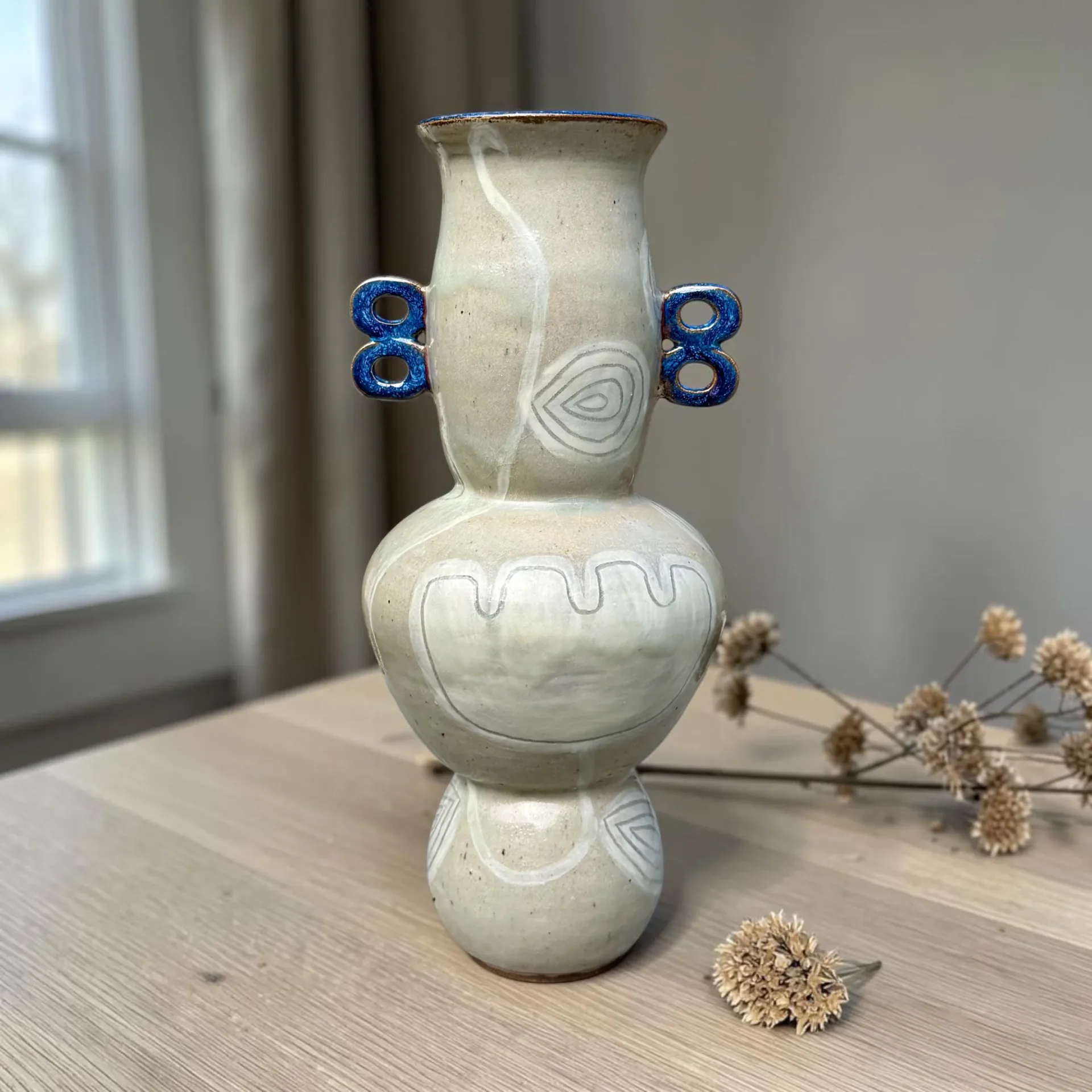 Pale celadon ceramic vase with bulbous form, cobalt blue handles, and incised linear patterns, displayed on wooden surface by Lisa Jakobsberg.