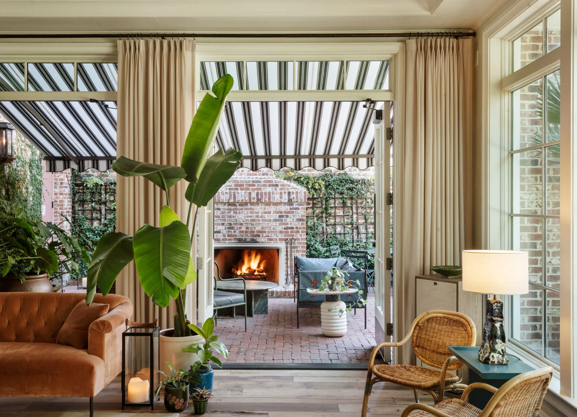 Open-concept interior with cognac leather sofa, woven chairs, and large potted banana plant opening onto brick patio with fireplace and pergola by Lisa Singer.