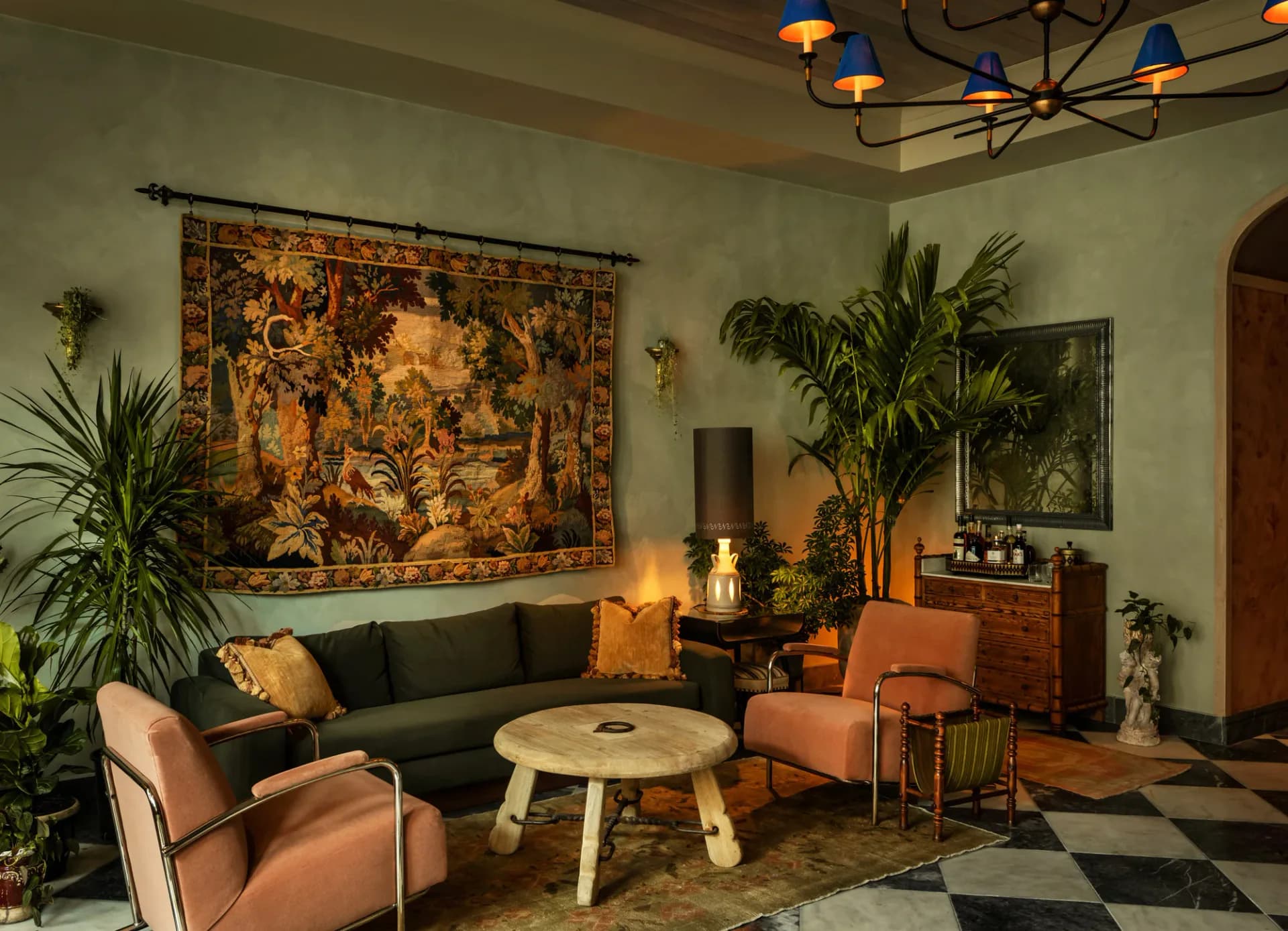 Warm living room interior featuring a dark green sofa, terracotta accent chairs, golden tapestry, abundant greenery, and elegant lighting by Lisa Singer.