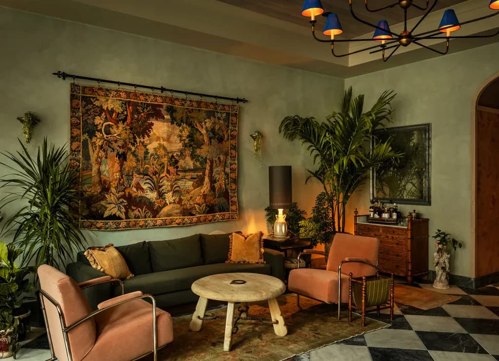 Warm-toned living room with sage green walls, terracotta seating, patterned tapestry, potted plants, and round wooden table by Lisa Singer.