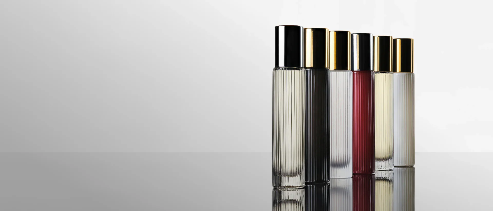 Glass cosmetic bottles with metallic caps arranged in a row, featuring clear and frosted glass in neutral and burgundy tones, product design by Lisa Sumner.