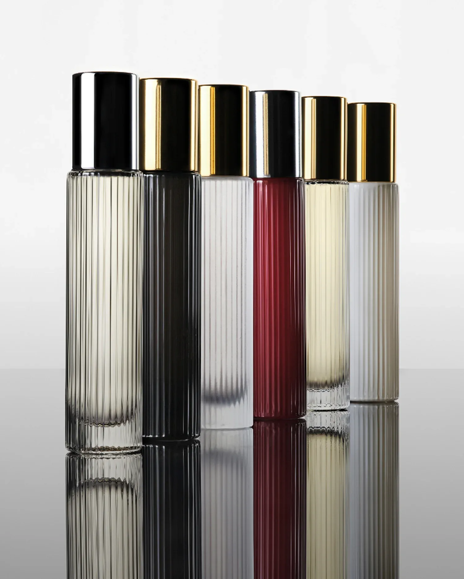 Six cylindrical ribbed glass bottles with black and gold caps in varying heights, filled with liquids ranging from clear to deep burgundy, arranged in a row by Lisa Sumner.
