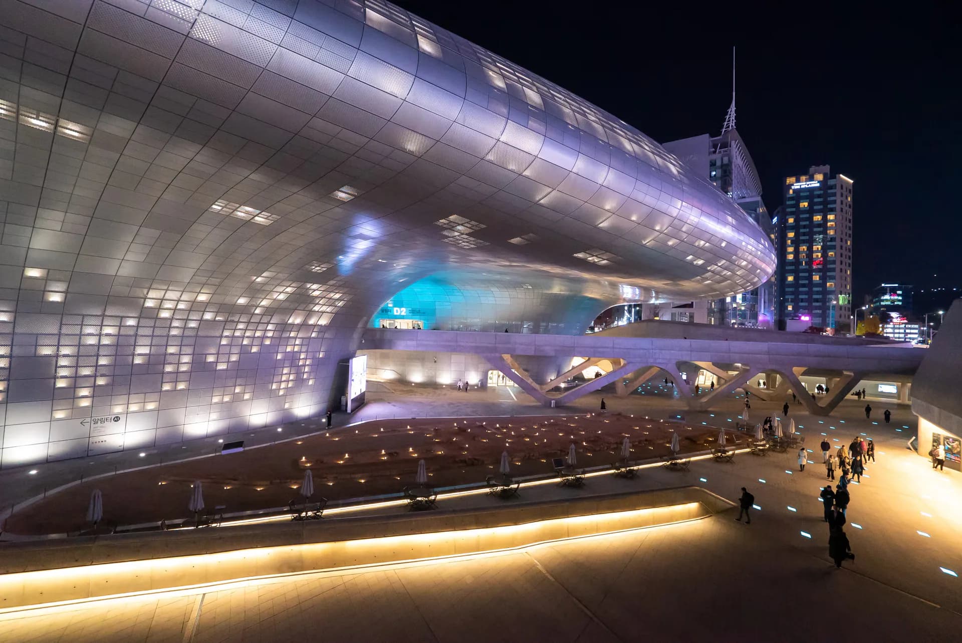 Illuminated modern architectural facade with pixelated silver paneling and curved forms at night, featuring warm ground-level lighting and urban skyline backdrop by Lisa-thi Beskar.
