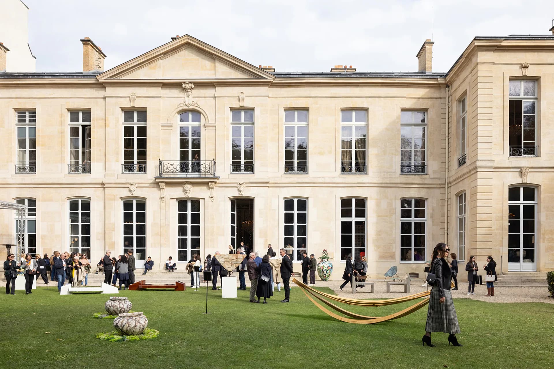 Neoclassical courtyard with cream-colored stone facades, tall arched windows, green lawn with sculptural installations, and guests gathered for an event by Lisa-thi Beskar.