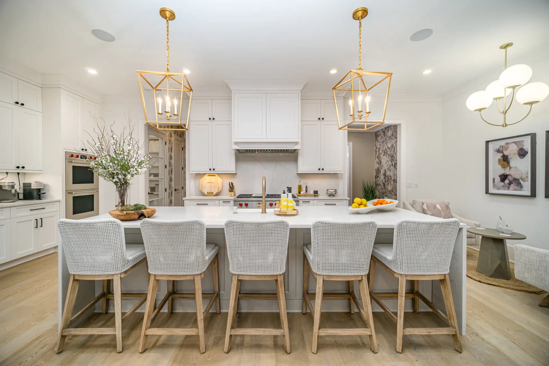 Bright modern kitchen with white cabinetry, marble countertops, and a large island with gray upholstered seating, brass pendant lighting, and warm wood flooring by Liz Aragona.