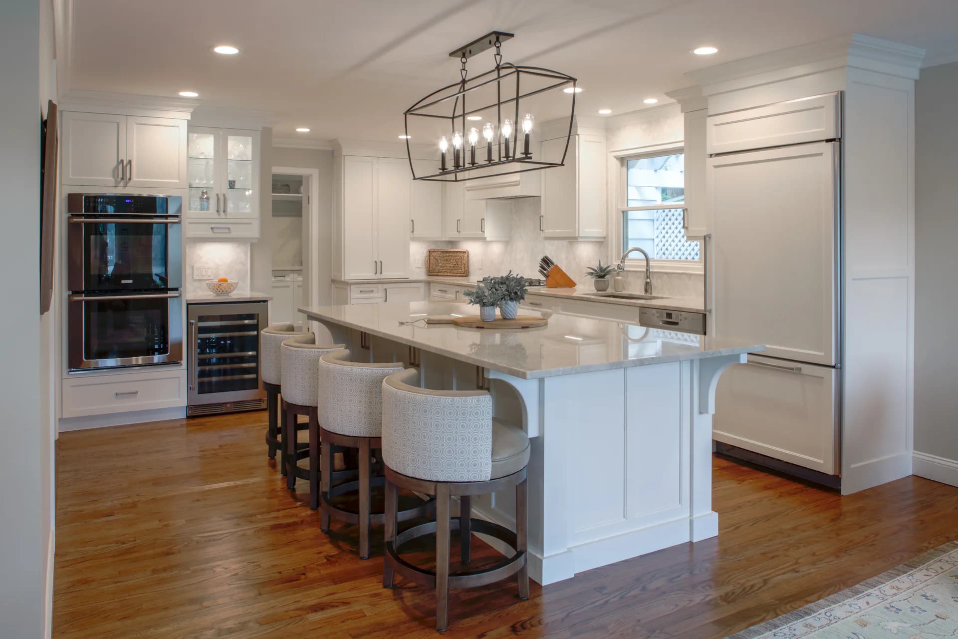 Modern white kitchen with large island, stainless steel appliances, wooden flooring, and pendant lighting fixture by Liz Aragona.