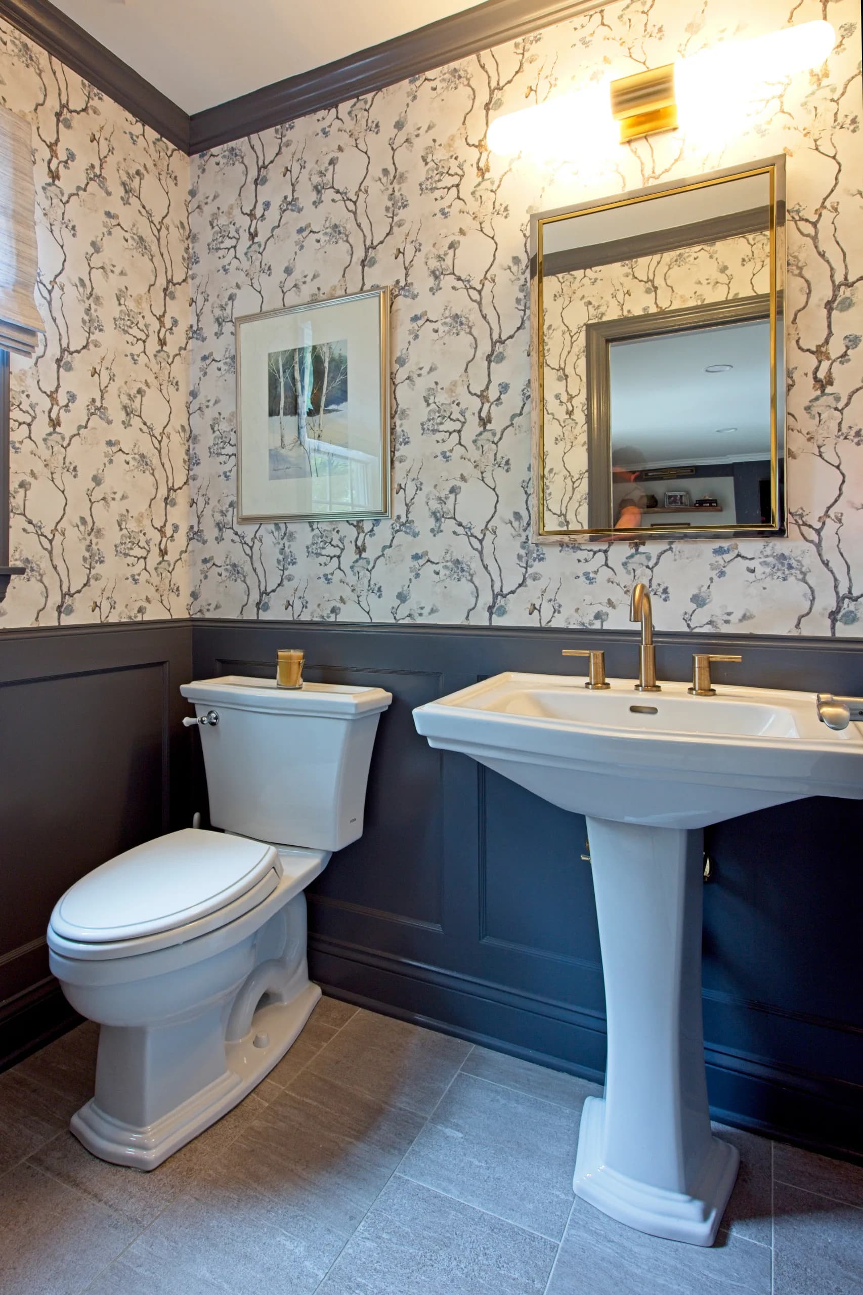 Elegant bathroom with cream and navy botanical wallpaper, pedestal sink with brass fixtures, framed mirror, and wainscoting detail by Liz Aragona.